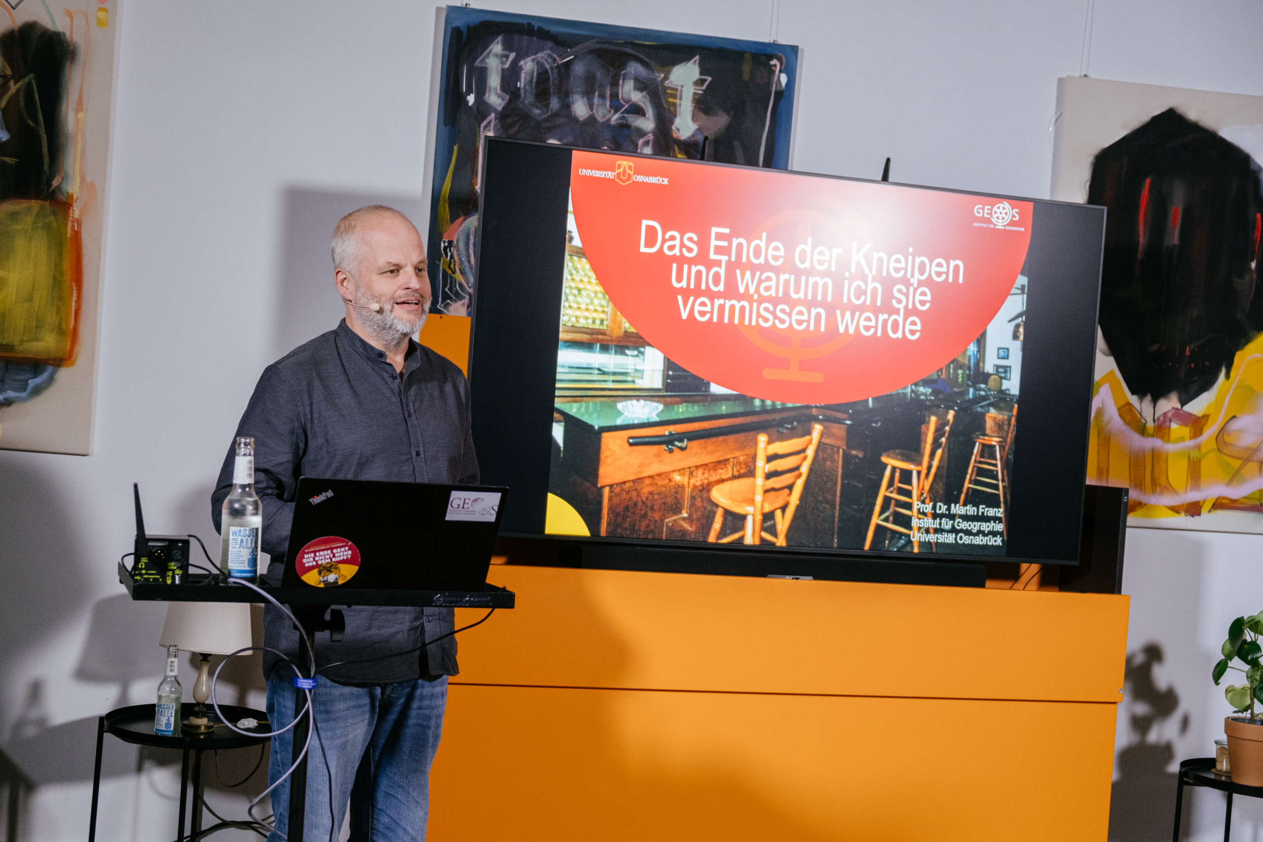 Prof. Dr. Martin Franz vor einem Bildschirm im Cafe Ütelier. Auf dem Bildschirm ist ein Foto einer Kneipe zu sehen und die Überschrift "Das Ende der Kneipen und warum ich sie vermissen werde:"